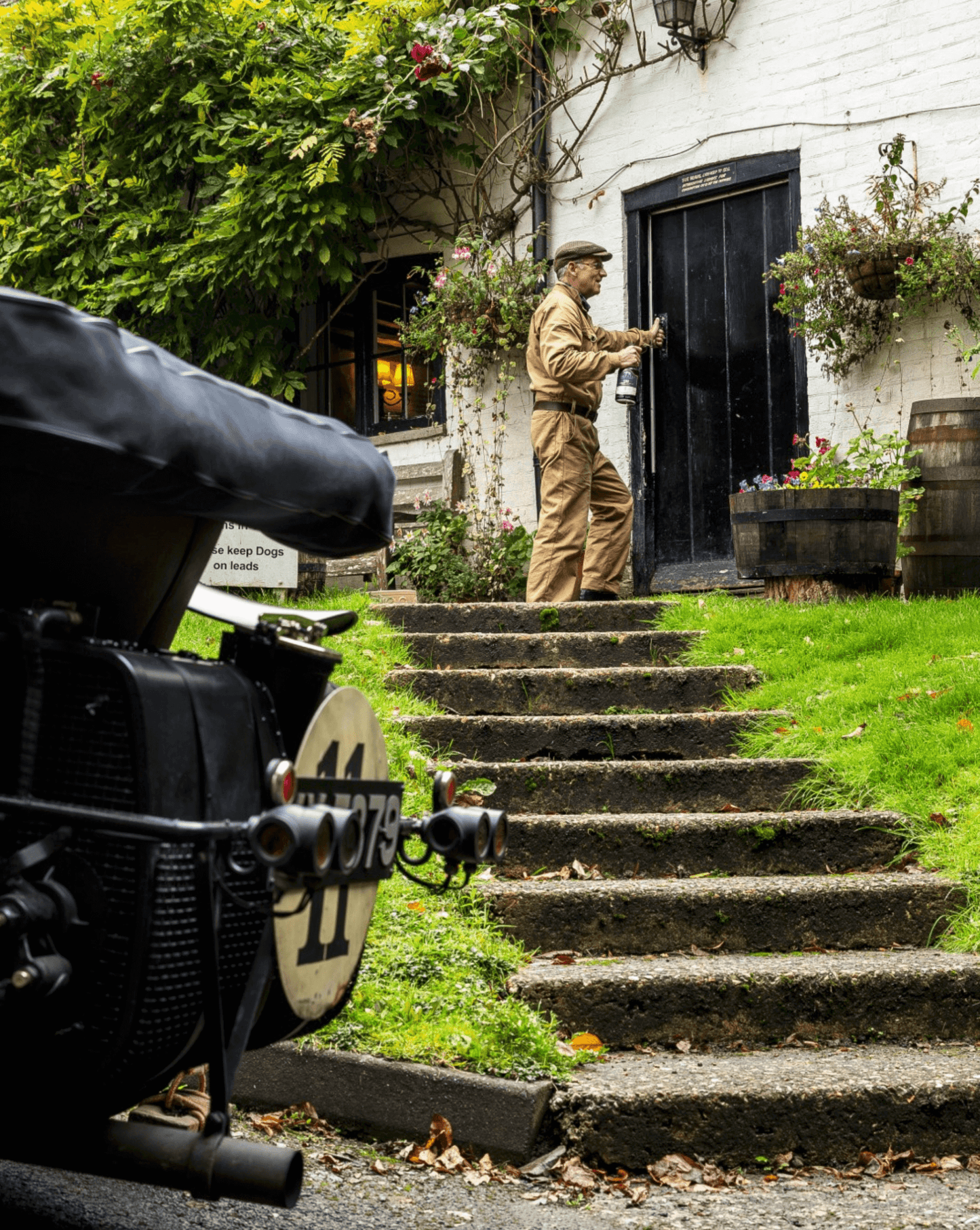 A man in vintage attire approaching a door with steps surrounded by greenery, showcasing a serene countryside scene.