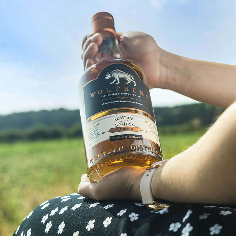 A hand holding a bottle of Aurora Single Malt Scotch Whisky in a scenic outdoor setting.