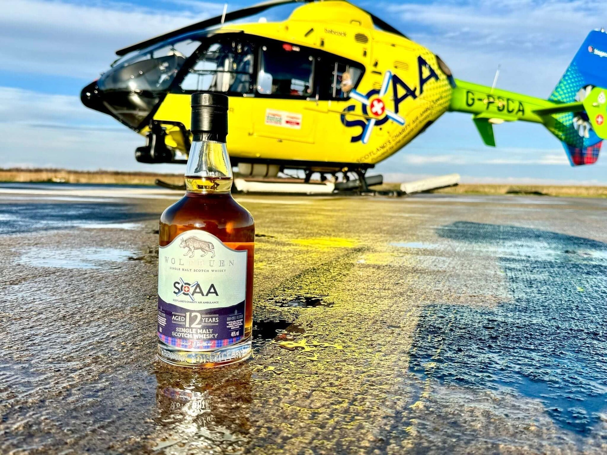 Scotland’s Charity Air Ambulance whisky bottle in front of a yellow helicopter on a runway.