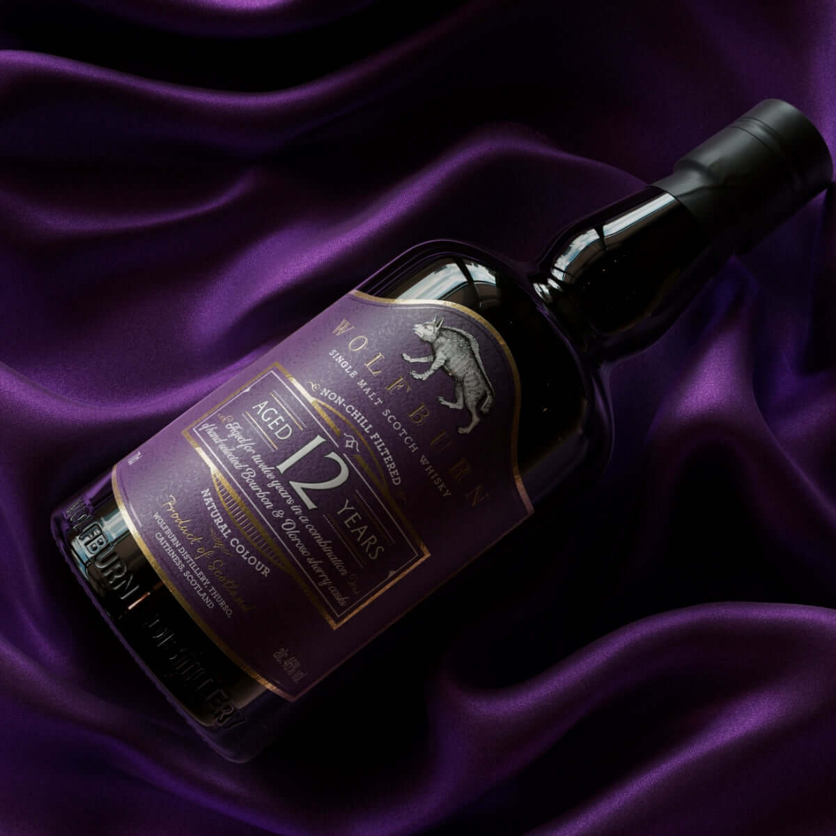 A bottle of Wolfburn 12-year-old single malt whisky lies on a smooth, purple satin fabric, with a purple and gold label prominently displayed.