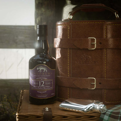 A bottle of 12-year-old Wolfburn single malt whisky sits on a wicker basket beside a leather case and a metal hip flask, evoking a rustic, elegant vibe.