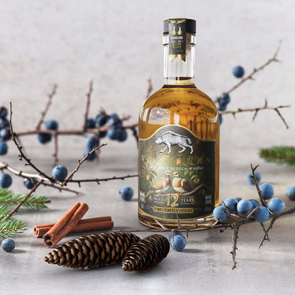 Bottle of whisky with a label featuring two turtle doves, surrounded by blueberries and cinnamon sticks on a light background.