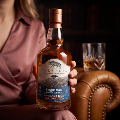 Woman holding a Wolfburn 12 Year Old Madeira Cask Finish bottle beside a leather armchair and a glass of whisky