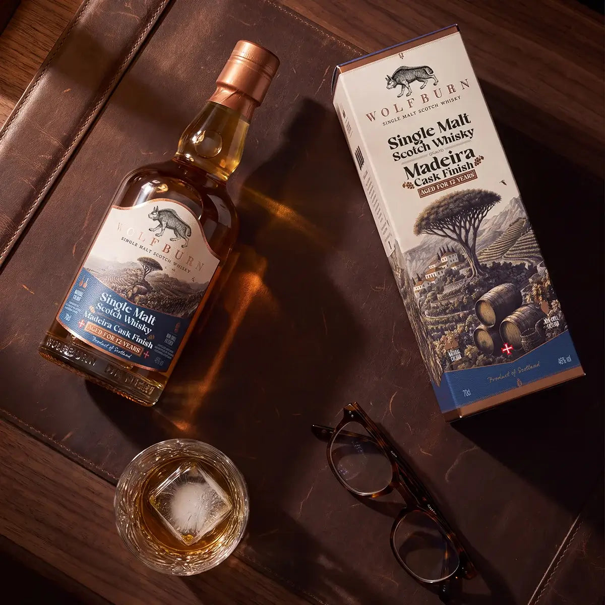 Wolfburn 12 Year Old Madeira Cask Finish bottle and carton on a leather desk with a glass of whisky over ice