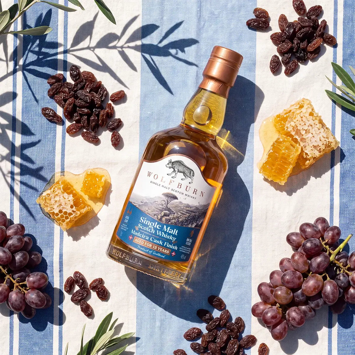 Wolfburn 12 Year Old Madeira Cask Finish bottle on striped linen with sultanas, honeycomb and grapes in sunlight