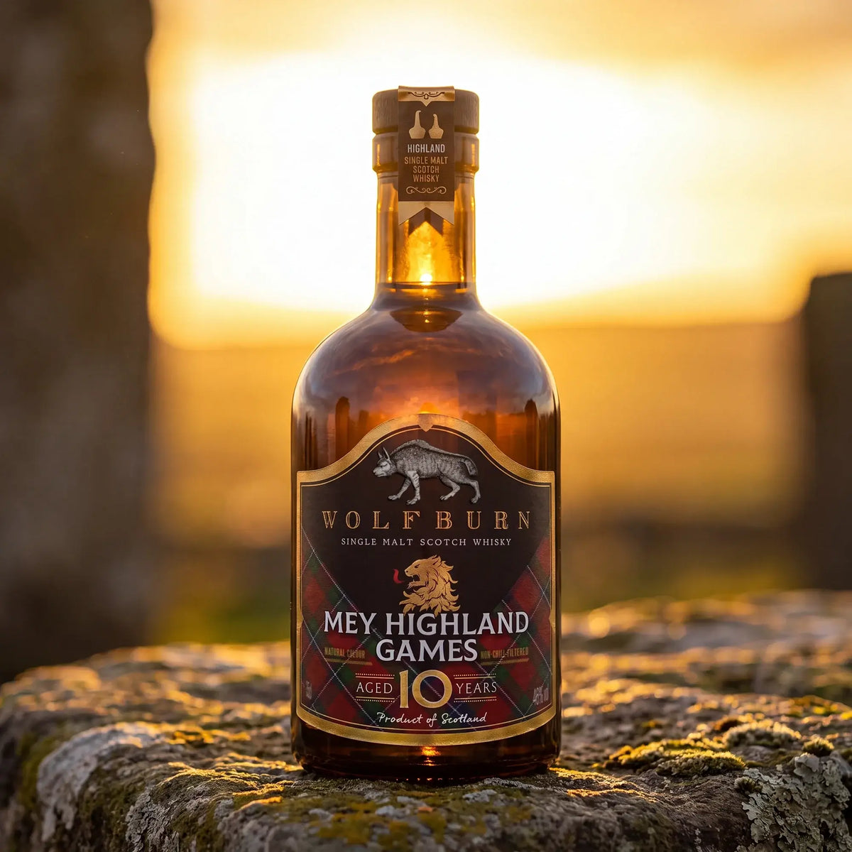 A bottle of Wolfburn 10 Years Old single malt Scotch whisky stands on a mossy stone at sunset, with a blurred outdoor landscape in the background. The label reads 'Mey Highland Games'.