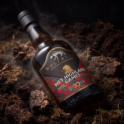 A bottle of Wolfburn 'Mey Highland Games' single malt scotch whisky lies on dark peat with visible smoke around it. The label features a red tartan pattern with gold and white text and graphics.