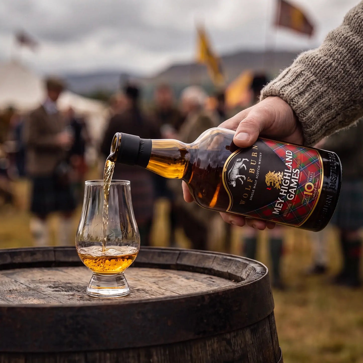 A hand pours amber whisky from a bottle labeled 'Mey Highland Games' into a small Glencairn glass on a wooden barrel, with a blurred outdoor event background.
