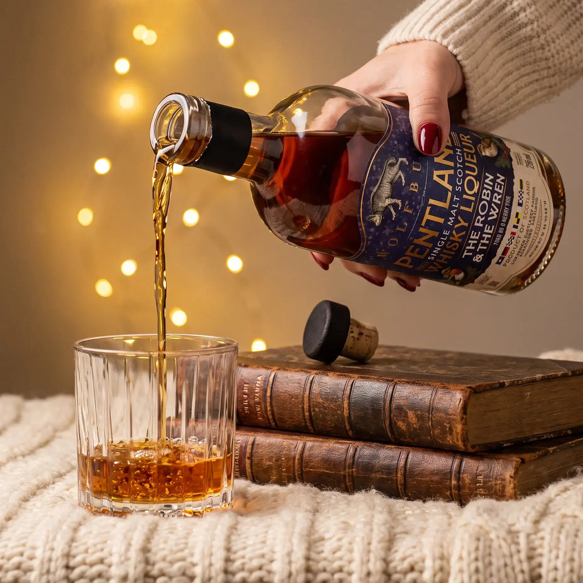 Pouring The Robin and The Wren Pentland Christmas whisky liqueur into a glass beside old books and fairy lights